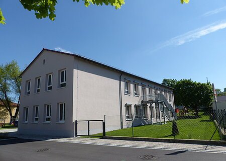 Kindergarten Monheim Städtischer Kindergarten in Monheim: ein zweistöckiges Gebäude mit weißer Fassade, Fenstern und Außentreppe, umgeben von einem Zaun und Bäumen. Rechts um das Gebäude ist ein Garten zu sehen. Vorderhalb des Gebäudes verläuft eine Straße.