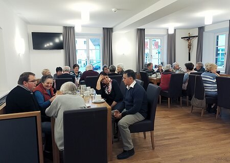 Bürgertreff "ZAMSEI" in Monheim - jeden 2. Samstag im Monat Der Bürgertreff "ZAMSEI" in Monheim: Viele Menschen sitzen bei Kaffee oder Tee in einem Raum an Tischen und unterhalten sich gesellig. Im Hintergrund sind drei Fenster mit Vorhängen und es hängt ein Jesus-Kreuz und ein Fernseher an der Wand.