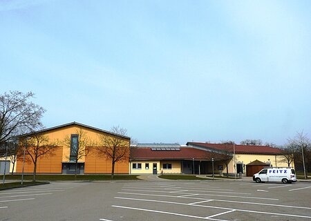 Stadt-/Mehrzweckhalle Monheim Stadt-/Mehrzweckhalle in Monheim: Ein großes, modernes Gebäude mit gelber Fassade und rotem Dach und mehreren Bäumen. Vorderhalb des Gebäudes ist ein großer Parkplatz, auf dem ein weißes Auto steht.