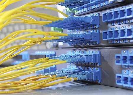 Breitbanderschließung Gelbe Glasfaserkabel sind in blaue Anschlüsse eines Netzwerkservers eingesteckt.