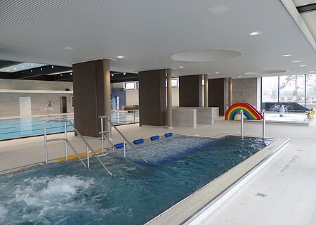 JURABAD Monheim Innenansicht eines Schwimmbads mit mehreren Becken, Rutsche in Regenbogenfarben und Wasserspielen.