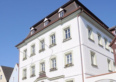 Das Rathaus der Drei-Stämme-Stadt Monheim Das Rathaus der Stadt Monheim, am Marktplatz. Ein weißes, dreistöckiges Gebäude mit Satteldach und mehreren Fenstern, im Vordergrund gepflasterter Marktplatz.
