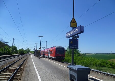 Bahnhof Otting-Weilheim Der Bahnhof Otting-Weilheim mit rotem Zug auf dem Gleis, blauer Himmel, grüne Landschaft im Hintergrund.