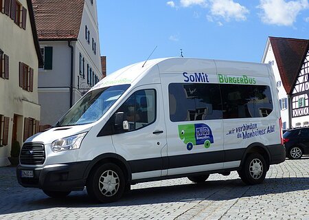 SoMit Bürgerbus Weißer Kleinbus steht auf einem gepflasterten Platz, in der Monheimer Innenstadt . Auf dem Bus ist die Aufschrift "SoMit BürgerBus" und "wir verbinden die Monheimer Alb".