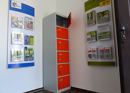 Indoor-Ladeschrank für eBike-Akkus und Infomaterial im Vorraum der Tourist-Information Monheim Ein orange-grauer Indoor-Ladeschrank für eBike-Akkus mit insgesamt sechs Fächern und einem offenen Fach steht zwischen zwei Wanddisplays mit Broschüren in dem Vorraum der Touristinformation Monheim.