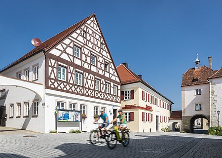Tourist-Information der Stadt Monheim/Monheimer Alb - im Schindlerhaus Tourist-Information der Stadt Monheim/Monheimer Alb: ein Fachwerkhaus mit drei Stockwerken, weißen Wänden und braunen Balken. Davor steht ein Schaukasten und zwei Radfahrer fahren auf gepflasterter Straße vorbei. Rechts davon ein gelbes zweistöckiges Gebäude mit roten Fensterläden.