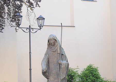 Statue der Hl. Walburga Monheim Steinstatue der heiligen Walburga in Monheim mit Stab, umgeben von Blumen in Töpfen und Laternen, vor einer hellen Gebäudewand, der Stadtpfarrkirche Monheim. Vor dem Gebäude sind Pflanzen eingesetzt.