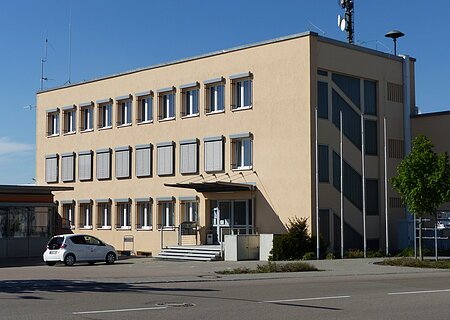 Caritas Sozial-Station Monheim Dreistöckiges Gebäude mit beigen Wänden, die Caritas Sozial-Station in Monheim, mit vielen Fenstern, einer Treppe vor dem Gebäude und einem geparkten Auto auf dem Parkplatz. Vorderhalb des Gebäudes verläuft eine Straße.