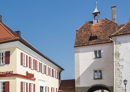 Tourist-Information der Stadt Monheim/Monheimer Alb - im Schindlerhaus Tourist-Information der Stadt Monheim/Monheimer Alb: ein Fachwerkhaus mit drei Stockwerken, weißen Wänden und braunen Balken. Davor steht ein Schaukasten und zwei Radfahrer fahren auf gepflasterter Straße vorbei. Rechts davon ein gelbes zweistöckiges Gebäude mit roten Fensterläden.