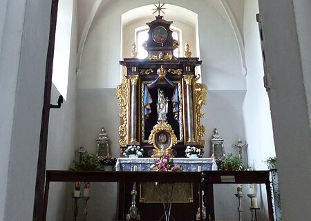 Walburgakapelle in der Stadtpfarrkirche St. Walburga Monheim Ein Blick durch eine Tür zu einem Kirchenaltar in der Walburgakapelle in Monheim, mit goldenen Verzierungen und einer Statue, der heiligen Walburga. Der Kirchenaltar ist umgeben von Kerzen und Blumen, in einem gewölbten Raum mit einer verzierten Decke.