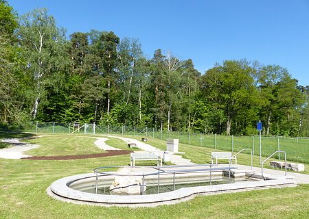 Kneipp Gesundheitsanlage Monheim mit Barfußpfad Monheim Rundes Kneippbecken in Monheim mit einem Geländer und einem Barfußpfad auf einer Wiese, umgeben von einer großen Grünfläche und Bäumen und Sitzmöglichkeiten. Ein Zaun im Hintergrund.