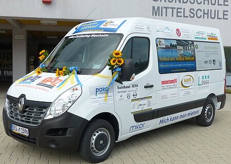 Das neue Carsharing-Modell in Monheim Ein weißer Transporter vom Car-Sharing-Anbieter Mikar mit Werbeaufdrucken und Sonnenblumen steht vor einem Gebäude mit der Aufschrift "Grundschule Mittelschule" in Monheim.