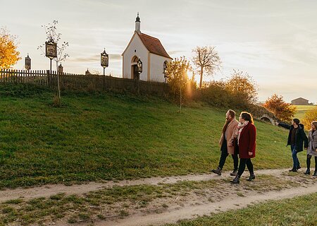 Die Kalvarienbergkapelle "Zur schmerzhaften Muttergottes" in Wittesheim - eingerahmt von 14 Kreuzwegstationen Vier Personen spazieren auf einem Weg an einer kleinen Kapelle vorbei, der Kalvarienbergkapelle in Wittesheim, umgeben von herbstlicher Landschaft. Auf dem Weg vor der Kapelle stehen Kreuzwegstationen.