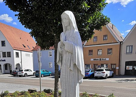 Marienstatue "Maria, Königin des Friedens" Statue einer betenden Frau mit Rosenkranz, die Marienstatue auf einem kleinen Podest, umgeben von Blumen, im Hintergrund eine Straße und ein Parkplatz mit Gebäuden.