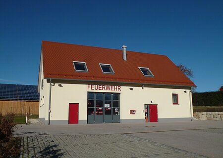 Feuerwehrhaus Weilheim Gelbes Feuerwehrgebäude mit rotem Dach, Aufschrift "Feuerwehr" an der Fassade, zwei rote Türen, ein großes Tor und Fenster auf der Vorderseite. Vorderhalb gepflasterter Platz und blauer Himmel im Hintergrund.
