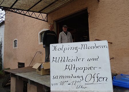 Kolpingsfamilie Altkleider- und Altpapiersammlung Zwei Personen stehen vor einem orangegestrichenen Gebäude neben einem Schild mit der Aufschrift "Kolping-Monheim Altkleider und Altpapier-Sammlung Offen".