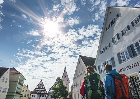 Wanderer in Monheim Zwei Personen mit Rucksäcken gehen durch eine Stadt mit Fachwerkhäusern unter einem sonnigen Himmel. Auf dem Haus im Vordergrund steht die Aufschrift "Stadtapotheke".