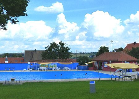 Freibad Monheim "Nostalgiebad" Freibad mit blauem Wasser, umgeben von Rasen, Bäumen und Gebäuden unter einem bewölkten Himmel. Vorderhalb vom Becken sind Sitzmöglichkeiten.