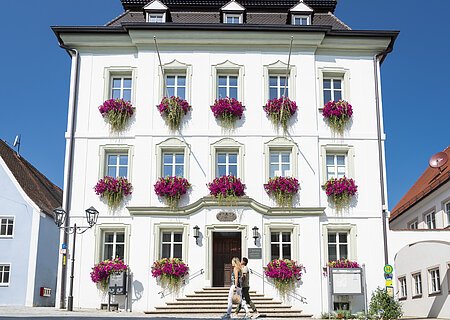 Das Rathaus der Drei-Stämme-Stadt Monheim Das Rathaus in Monheim: Ein weißes dreistöckiges Gebäude mit mehreren Fenstern, an denen Blumenkästen mit pinken Blumen hängen. Zwei Personen gehen davor vorbei. Links und rechts davon jeweils ein weiteres Haus.