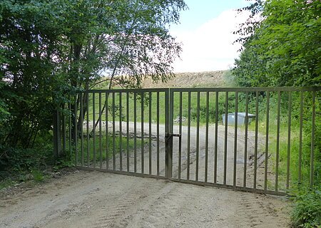 Erdaushubdeponie Monheim Verschlossenes Metalltor auf einem unbefestigten Weg, umgeben von Bäumen und Gras. Im Hintergrund ist ein Müllcontainer und ein Hügel sichtbar.
