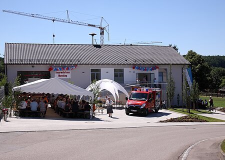 Neues Feuerwehrhaus Warching, Schloßberg 4 Feuerwehrhaus mit einem weißen Festzelt, einem weißen Pavillon und einem Feuerwehrfahrzeug davor. Menschen sitzen unter dem Zelt, ein Kran steht hinter dem Feuerwehrhaus, im Vordergrund eine gepflasterte Straße. Feuerwehrhaus ist umgeben von Feldern und einem Wald.