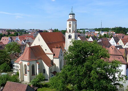 Stadtpfarrkirche St. Walburga Monheim Luftbild der Stadtpfarrkirche mit Turm in einer Stadtlandschaft, umgeben von Häusern mit roten Dächern und Bäumen. Im Hintergrund blauer Himmel.