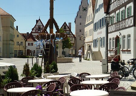 Café Konditorei Wenninger - das Café am Marktplatz Ein Marktplatz mit Tischen und Stühlen eines Cafés im Vordergrund, Fachwerkhäusern im Hintergrund und einem Maibaum in der Mitte des Marktplatzes. Eine Person läuft auf dem Markplatz.