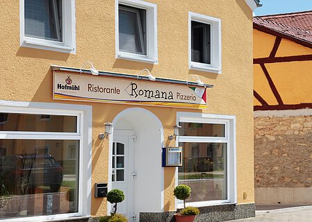 Pizzeria Ristorante Romana - Mediterraner Charme in Monheim Gelbes Gebäude mit mehreren Fenstern und einem Schild "Ristorante Romana Pizzeria" über dem Eingang. Zwei kleine Bäume links und rechts vor der Tür.