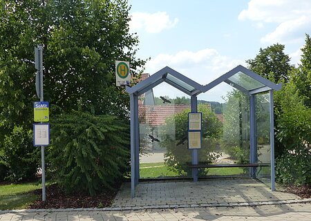 Bushaltestelle Weilheim Bushaltestelle mit Glasüberdachung, umgeben von Bäumen und Sträuchern, mit Fahrplan und Haltestellenschild.