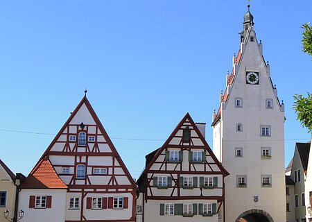 Oberer Torturm mit Moserhäusern - Blick aus der Innenstadt Historische Fachwerkhäuser, ein Torturm mit Uhr in einer Altstadt bei klarem Himmel. Links im Bild parkende Autos auf einem gepflasterten Platz. Rechts im Vordergrund ein Baum mit einer Info-Tafel und einem großen Buchstaben "O" aus Stein. Dahinter eine Informationsvetrine.