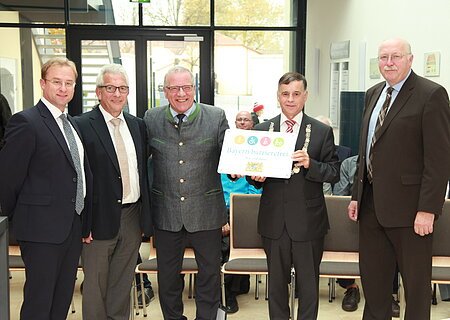 "Bayern barrierefrei" Fünf Männer in Anzügen stehen in einem Raum, einer hält ein Schild mit der Aufschrift "Bayern barrierefrei". Im Hintergrund große Eingangstür aus Glas.