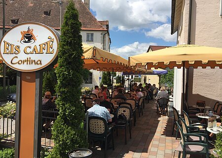 Eiscafé Cortina Außenbereich eines Eiscafés mit gelben Sonnenschirmen, Tischen und Stühlen, Gäste sitzen und essen.