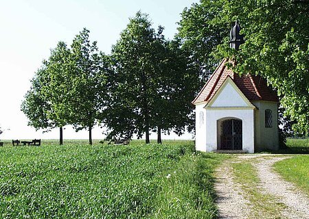 Die Brandkapelle in Monheim Kleine Kapelle mit rotem Dach auf einer Wiese mit einem Schotterweg. Umgeben von einem grünem Feld und Bäumen.