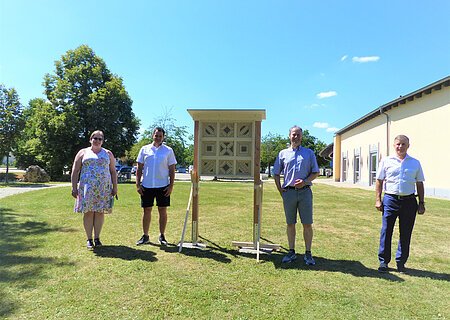 Wildbienen Nisthilfe Vier Personen stehen auf einer Wiese links und rechts neben einem großen Insektenhotel, im Hintergrund Bäume, blauer Himmel und ein weiß gestrichenes Gebäude.