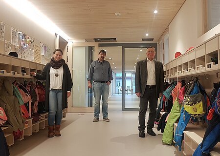 Über den neuen Kindergarten freuen sich die Leiterin Susanne Utjesinovic, Stadtbaumeister Richard Meyer und Bürgermeister Günther Pfefferer (v.l.n.r) Drei Personen stehen in einem Flur mit Kindergarderoben und bunten Jacken. Im Hintergrund ist ein Raum mit Tischen und kleinen Stühlen sichtbar.