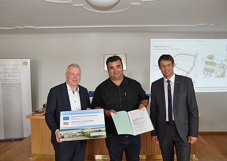 ELER Förderbescheid erhalten für das Projekt "Dorferneuerung - Straßenausbau Flotzheim" Drei Männer stehen in einem Raum, einer hält ein Plakat, ein anderer ein Dokument in den Händen. Im Hintergrund ein Banner vom Amt für Ländliche Entwicklung in Bayern und eine Präsentation an der Wand mit einer Karte.