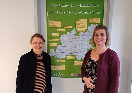 LAG Geschäftsstelle Monheim Zwei lächelnde Personen stehen vor einem grünen Plakat mit einer Karte der Region Monheimer Alb-AltmühlJura. Umgeben von einer weißen Wand.