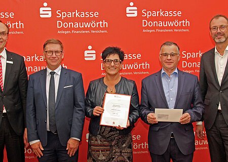 Monheim summt - Nachhaltigkeitspreis 2023 Fünf Personen stehen vor einem roten Hintergrund mit dem Logo der Sparkasse Donauwörth. Eine Person hält ein Zertifikat und eine weitere Person einen Brief in den Händen.
