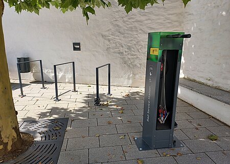 Rad-Service-Station und Fahrradabstellanlagen Stadt Monheim Fahrradservice-Station mit Werkzeugen und Fahrradständern auf einem gepflasterten Gehweg unter einem Baum. Umgeben von einer weißen Mauer.