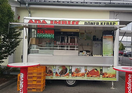 ADA Imbiss Döner-Kebab Monheim Imbisswagen mit geöffnetem Verkaufsfenster und zwei Stehtischen davor. Auf dem Wagen oben die Beschriftung „ADA Imbiss - Döner Kebab“ sichtbar unter dem geöffnetem Fenster verschiedene Abbildungen der erhältlichen Gerichte abgebildet. Links im Bild ein Baum, rechts ein Teil eines Parkplatzes zu sehen.