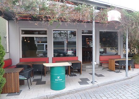 Vis-á-Vis - die kleine Kneipe für Jung und Alt Außenbereich einer Kneipe mit roten Sitzbänken, Holztischen und schwarzen Stühlen. Vor dem Gebäude ein grünes Fass als Tisch auf einem gepflastertem Platz aufgestellt. Am Dach entlang verläuft ein grünes Gewächs. Die Überdachung ist mit rot-gelben LED-Streifen beleuchtet.