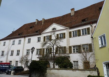 Ehemaliges Schloss / Amtsgericht Monheim Altes, historisches, mehrstöckiges Gebäude mit braunem Ziegeldach, weißen Wänden und grünen Fensterläden bei Tageslicht. Vor dem Gebäude sind Bäume, parkende Autos und eine Straßenlaterne zu sehen. Rechts im Bild Stück einer weißer Mauer und ein grünes Gebäude zu sehen.