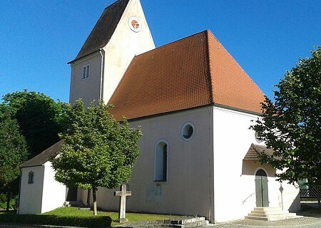 Filialkirche St. Johannes d. T. Rehau Kirche mit rotem Satteldach und einer Turmuhr umgeben von Bäumen bei klarem Himmel, an einem gepflastertem Weg.