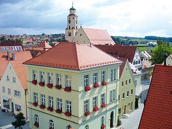 Haus des Gastes und im Hintergrund die Stadtpfarrkirche Monheim Blick von oben auf eine Innenstadt mit Hauptaugenmerk auf ein historisches grünes Gebäude im Vordergrund, mit roten Dachziegeln und Blumen an den Fenstern. Im Hintergrund weitere Häuser der Kleinstadt und blauer Himmel. Rechts unten im Eck das rote Dach eines anderen Gebäudes im Vordergrund zu sehen.