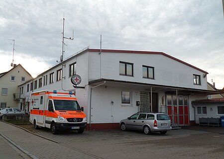 Bayerisches Rotes Kreuz - Rettungswache Monheim An der Straße gelegenes weißes Gebäude mit Deutschem Roten Kreuz-Schild seitlich am Gebäude, roten Toren und einem Rettungswagen links am Gebäude. Auf dem gepflastertem Platz steht ein grauer Kombi vor dem Gebäude, bei bewölktem Himmel.