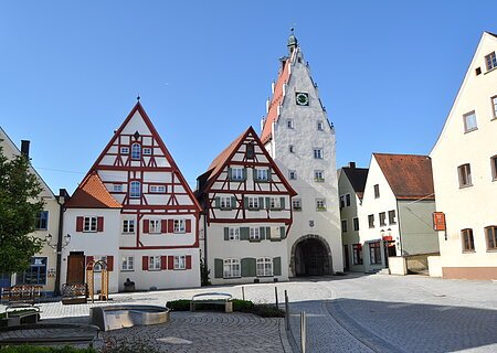 Moserhäuser mit dem Oberen Torturm Historische Fachwerkhäuser in einer Innenstadt mit einem weißen Uhrenturm an einem gepflasterten Platz bei klarem Himmel. Im Vordergrund ist ein Brunnen zu sehen, umgeben von drei Sitzbänken.