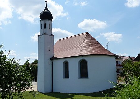 Filialkirche St. Apollonia Kölburg Weißes Kirchgebäude mit abgerundetem Grundriss und kleinem Turm mit schwarzer Haube bei blauem Himmel, umgeben von grünen Bäumen und wiese. Im Hintergrund weitere Häuser.