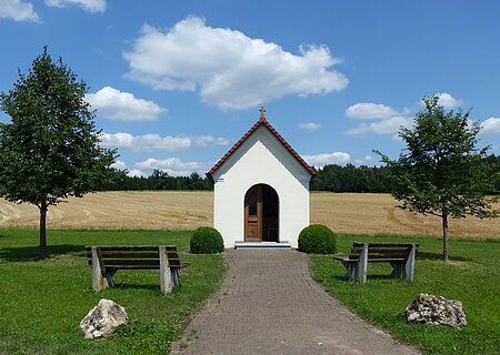 Kapelle Kreut Kleine weiße Kapelle mit rotem Dach an einem gepflastertem Weg mit jeweils einer Bank, einem Baum und einem großen Stein links und rechts am Weg entlang, auf einer Wiese vor Feld und blauem Himmel.