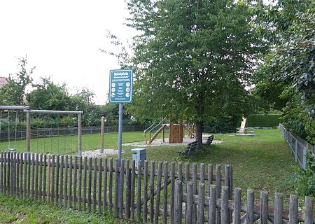 Spielplatz Flotzheim Spielplatz mit einem Holzzaun, Schaukeln, Klettergerüst, Rutsche, Bänken und Bäumen auf Grasfläche und teilweise mit Kieselsteinen bei Tageslicht. Außerdem ein am Zaun angebrachtes grünes Schild mit Regeln zur Spielplatzbenutzung und eine Mülltonne aus Metall zu sehen.