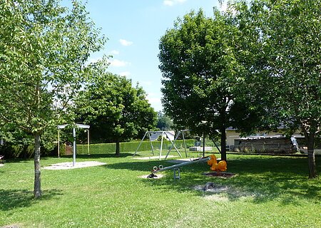 Spielplatz Itzing Spielplatz mit Wippen, Schaukeln und einem Sandkasten umgeben von grünen Bäumen auf einer Wiese bei Sonnenschein.